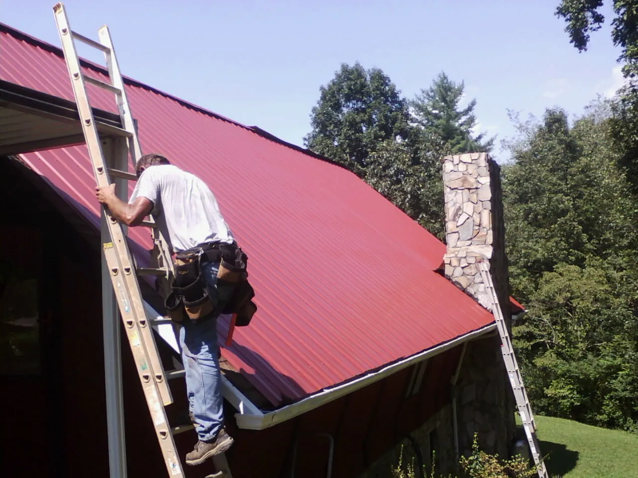 Roofer on a ladder beside a red metal roof and stone chimney — Soffit & Fascia Repair by Roofing Brothers in Lairdsville